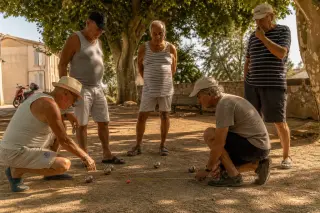Group of pétanque players chatting around a cochonnet on a shaded Provençal square with plane trees, metallic boules and measuring tape visible on the dusty ground in warm afternoon light.